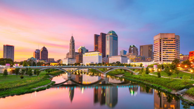 Downtown city skyline with modern buildings, a bridge, and river at sunset; sky glows with vibrant pink, orange, and blue colors, reflected in the calm water below.