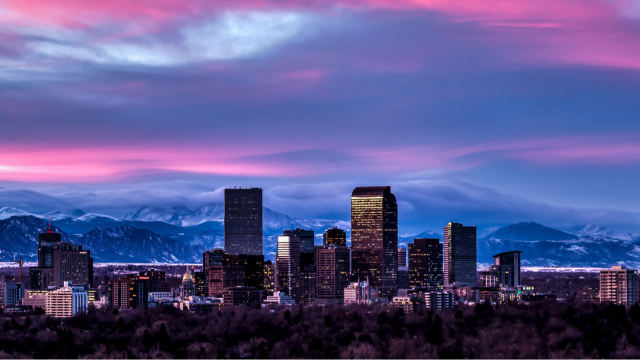 Denver city skyline at dusk with tall buildings silhouetted against a dramatic sky of pink, purple, and blue hues. Snow-capped Rocky Mountains are visible in the background.
