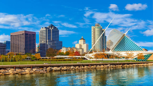 Downtown cityscape of Milwaukee, Wisconsin, featuring modern high-rise buildings, the Milwaukee Art Museum with its unique white architecture, trees with autumn foliage, and a waterfront under a blue sky with scattered clouds.