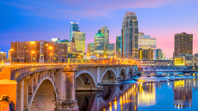 A city skyline at sunset with tall buildings lit up, a stone arch bridge over a river, and reflections of lights shimmering on the water.