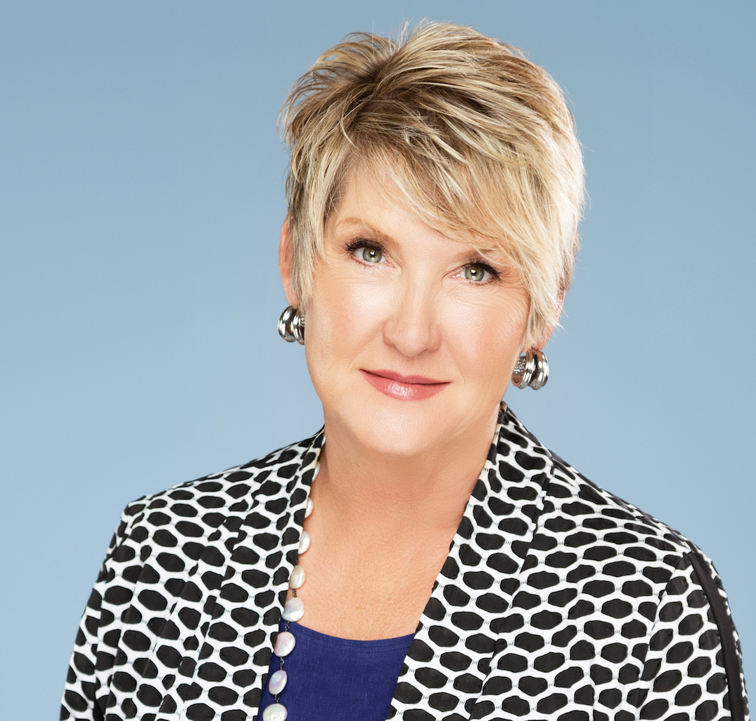 A middle-aged woman with short blonde hair wears a patterned black and white blazer over a blue top.