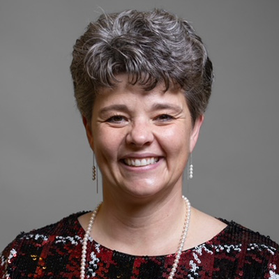 A woman with short, curly gray hair smiles at the camera. She is wearing a patterned top, a pearl necklace, and drop earrings. The background is a plain, gray backdrop.