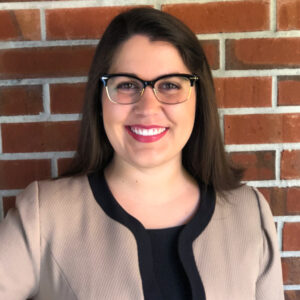 A smiling woman wearing glasses and a beige blazer against a brick wall.