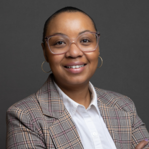 A smiling person with short hair, wearing clear-framed glasses and a plaid blazer over a white collared shirt.
