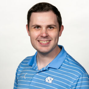 A smiling man wearing a light blue polo shirt with horizontal white stripes and the University of North Carolina logo.
