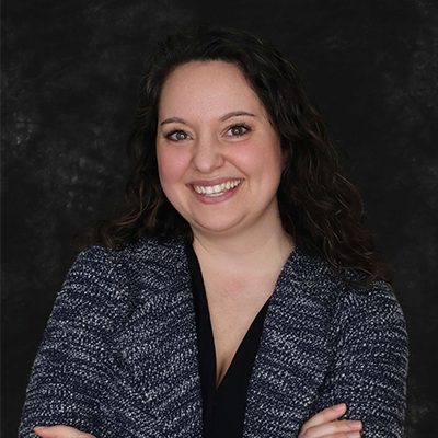 A woman with long curly dark hair smiles with her arms crossed, wearing a dark patterned cardigan over a black top, in front of a dark, textured background.
