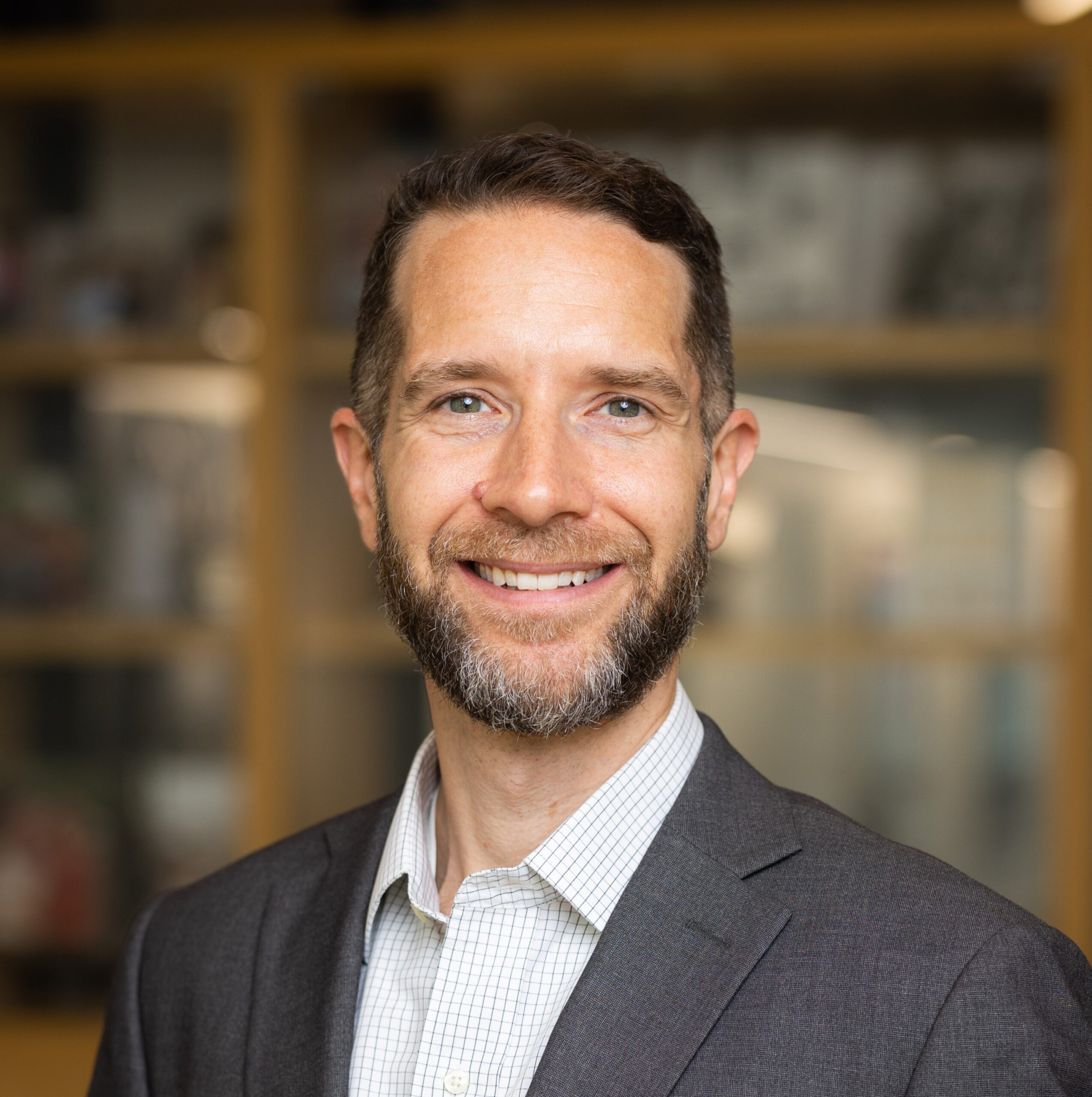 A man with short brown hair and a beard smiles at the camera. He is wearing a gray suit jacket over a light-colored checked shirt, standing indoors with blurred shelves in the background.