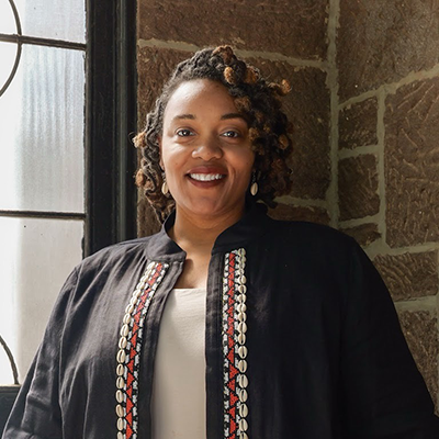 A woman with curly hair wearing a black jacket with red and white patterns stands smiling in front of a window and a stone wall.