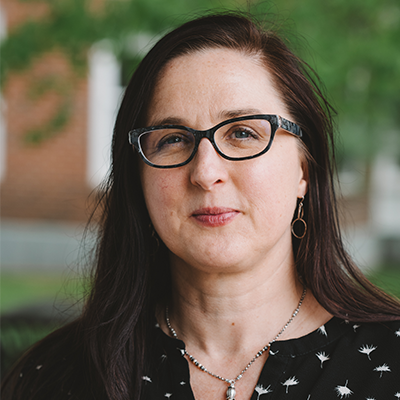 A woman with long brown hair, wearing glasses, a black patterned blouse, and a necklace, is standing outdoors with greenery and a brick building in the blurred background.
