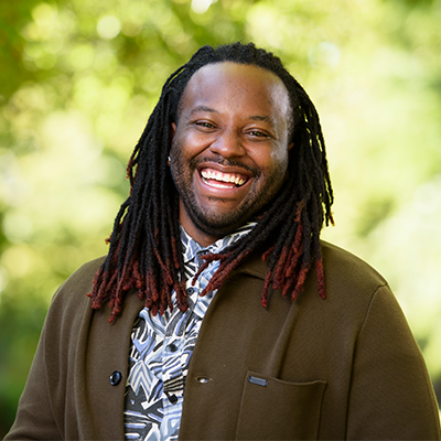 A man with long, dark dreadlocks and a bit of red at the ends smiles brightly. He wears a patterned shirt and a brown coat, standing outdoors with a blurred green background.