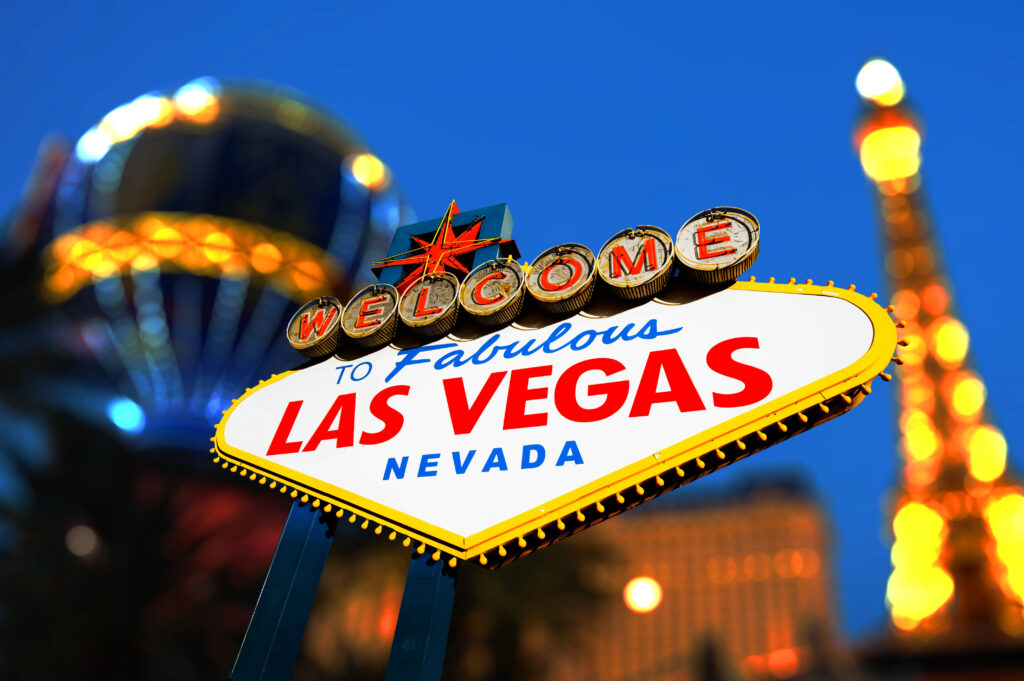 A close-up of the iconic Welcome to Fabulous Las Vegas, Nevada sign is shown brightly lit at night, with blurred city lights and landmarks in the background.
