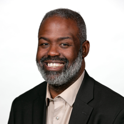 A smiling man with short gray hair and a beard, wearing a black blazer over a beige shirt, poses against a plain white background.