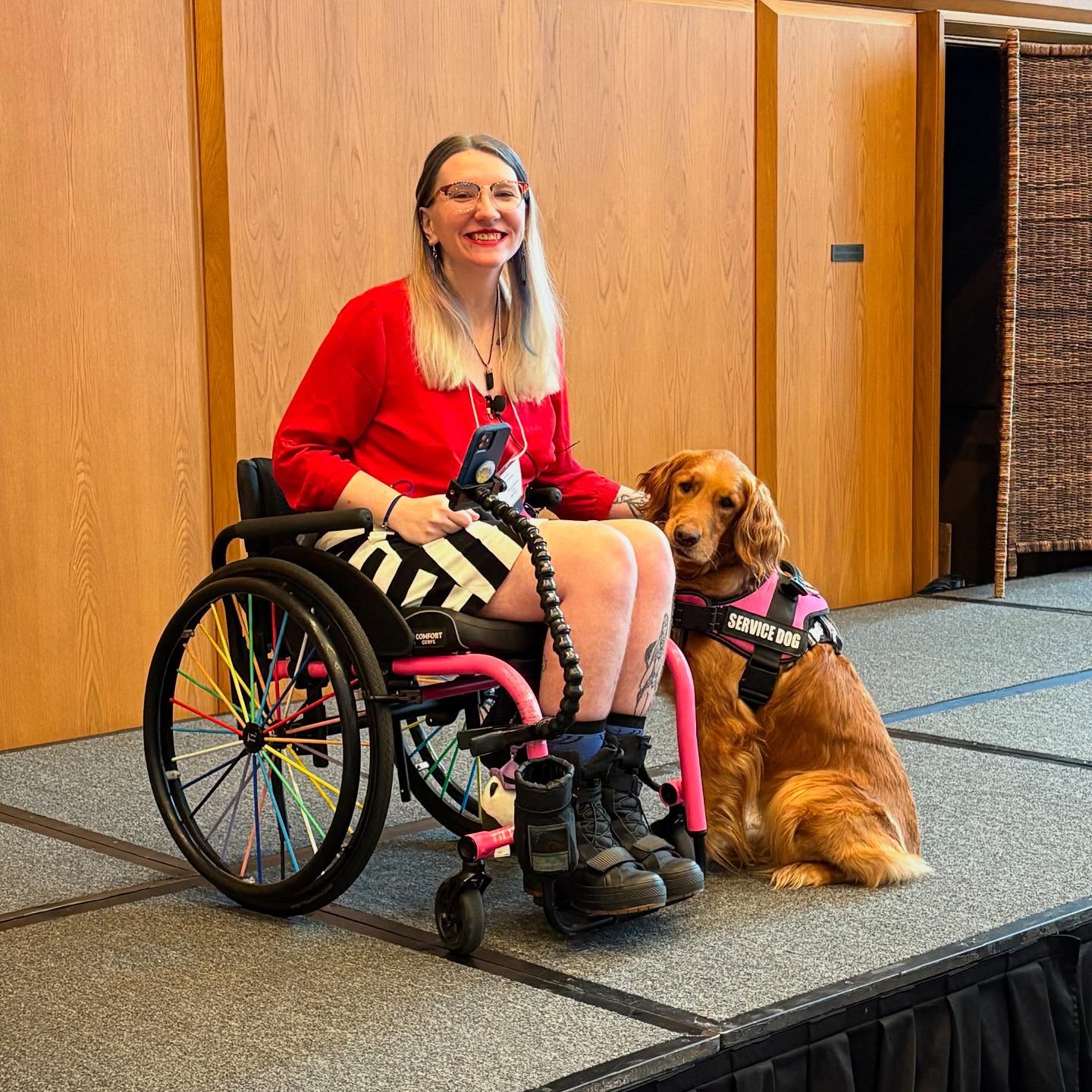 A smiling woman with long blonde hair, wearing a red top and black-and-white skirt, sits in a colorful wheelchair. A golden service dog in a pink vest sits beside her. They are on a stage with wooden paneling behind them.