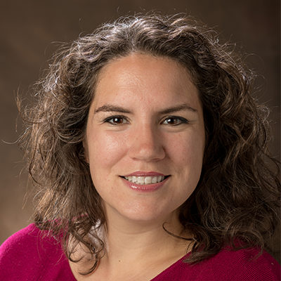 A woman with curly brown hair, wearing a pink top, smiles in front of a brown background.