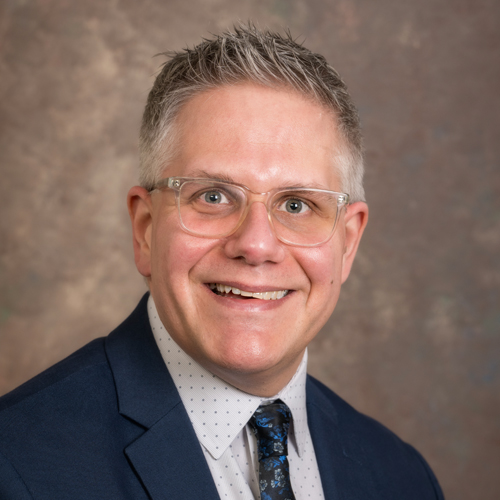 Smiling man with short gray hair wears clear glasses, a navy suit, a white shirt with small dots, and a patterned tie. He poses in front of a mottled brown and tan backdrop.