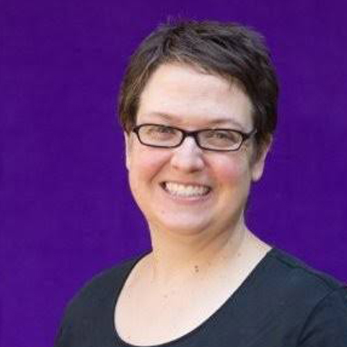 A smiling person with short brown hair and glasses, wearing a black top, stands in front of a solid purple background.