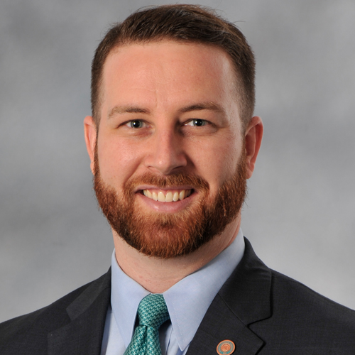 A man with short brown hair and a reddish beard, wearing a dark suit, light blue shirt, and teal tie, smiling in front of a gray background.