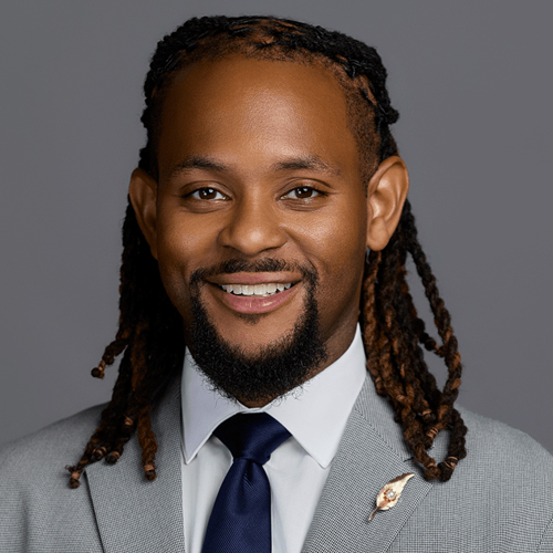 A smiling man with long, brown-tipped dreadlocks, a short beard, wearing a light gray suit, white shirt, and navy tie, posing in front of a plain gray background.