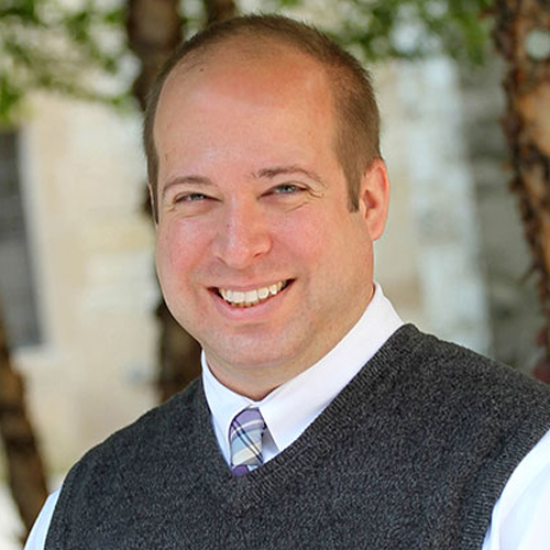 A man with short hair, wearing a white shirt, striped tie, and gray sweater vest, smiling outdoors with trees and a blurred background.