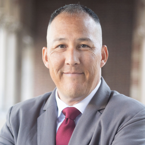 A man in a gray suit and red tie smiles at the camera with a blurred building background.