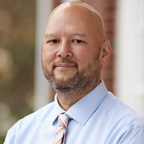 A man with a bald head and trimmed beard, wearing a light blue shirt and striped tie, stands outside in front of a blurred brick building background, looking calmly at the camera.