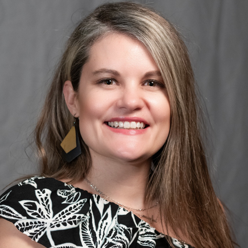 Smiling woman with long brown hair wearing a black and white patterned top, large geometric earrings, and a necklace, posing in front of a neutral gray background.
