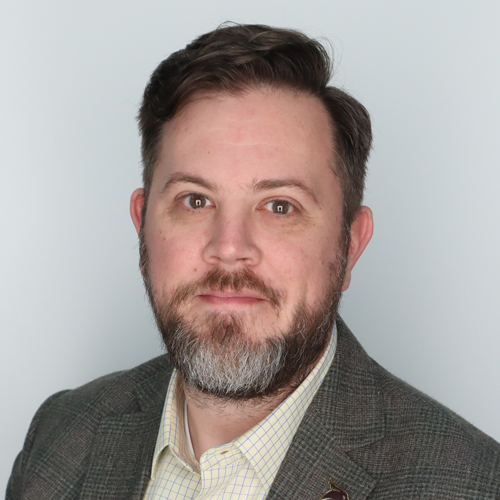 A man with short brown hair and a beard wears a gray blazer over a light-colored checkered shirt, posing in front of a plain light gray background.