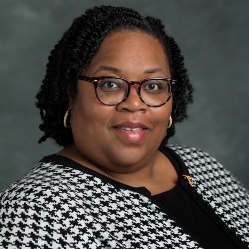 A woman with short, curly hair and glasses smiles at the camera. She is wearing a black and white houndstooth jacket over a black top, with gold hoop earrings, against a gray background.