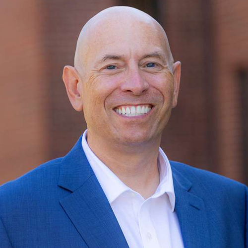 A smiling bald man in a blue suit jacket and white shirt stands outdoors in front of a blurred brick background.