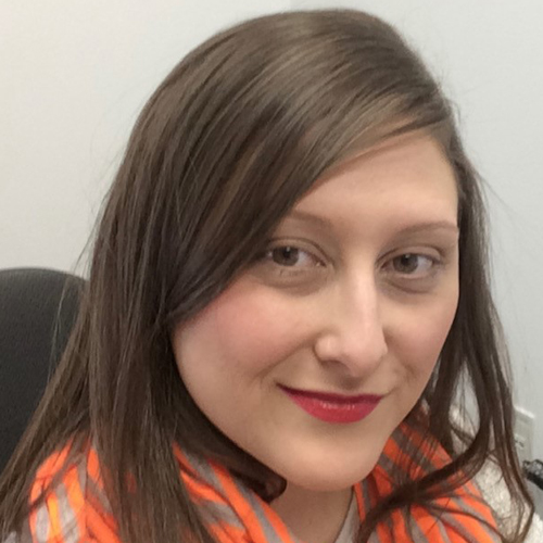 A woman with straight brown hair and red lipstick smiles slightly at the camera. She is wearing an orange and gray striped scarf and sits in an office chair against a light-colored wall.