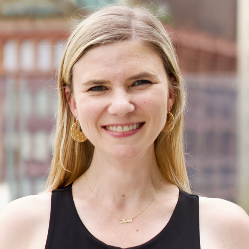 Smiling woman with straight blonde hair, wearing a black top, gold hoop earrings, and a gold necklace, standing outside with blurred buildings in the background.