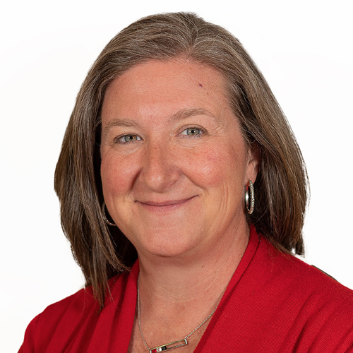 Smiling woman with straight, shoulder-length brown hair wearing a red top and silver hoop earrings, posed against a plain white background.