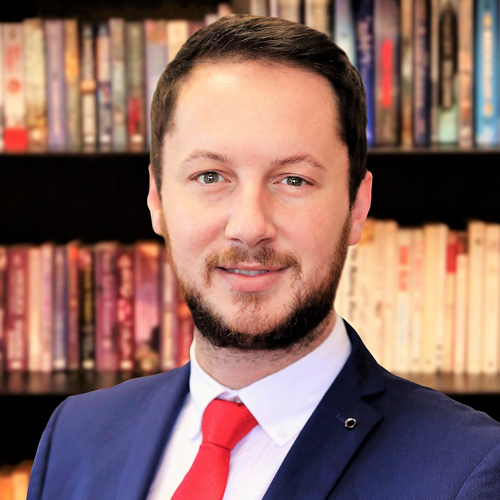 A man with short brown hair and a beard, wearing a blue suit, white shirt, and red tie, stands smiling in front of a blurred bookshelf filled with books.