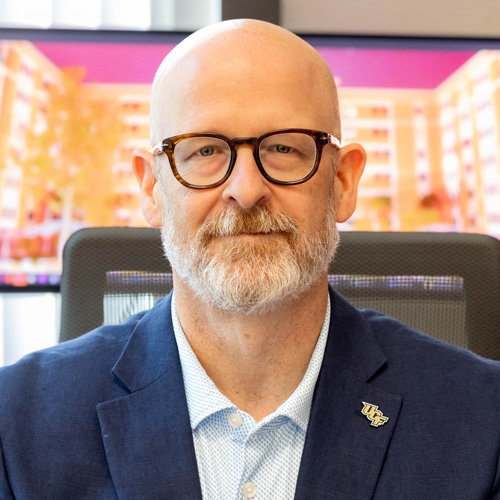 Bald man with glasses and a gray beard wearing a blue blazer and light shirt, sitting in an office chair. Two blurred screens in the background show colorful building images.