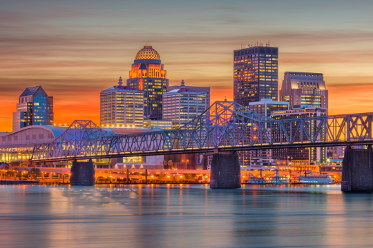 Louisville, Kentucky skyline at sunset with colorful sky, illuminated buildings, and a bridge crossing over calm water in the foreground.
