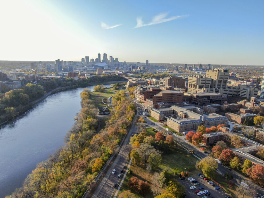 Aerial view of a city skyline with a river, autumn trees, and buildings, taken on a sunny day. Cars are parked along the road, and the city rises in the background.