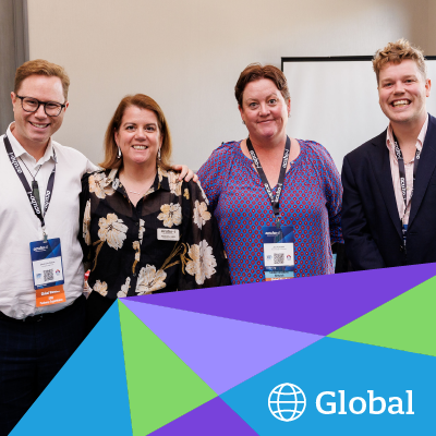 Four people wearing event lanyards pose for a group photo in front of a blue and purple geometric graphic with the word Global and a globe icon.