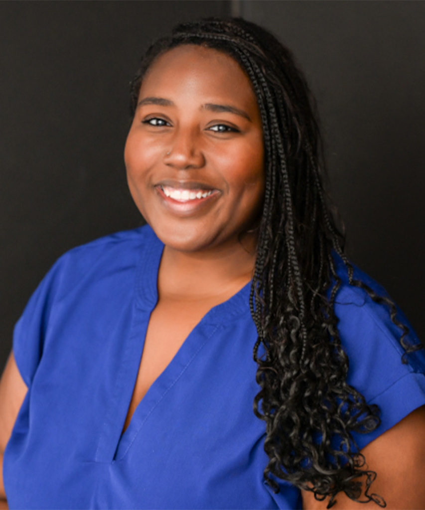 A woman with long braided hair smiles at the camera. She is wearing a blue V-neck top and stands against a dark background.
