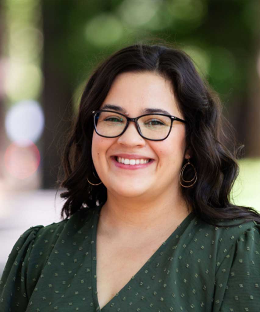 A woman with dark wavy hair, glasses, and hoop earrings smiles outdoors. She wears a green dress with small white dots. Blurred trees and sunlight are in the background.