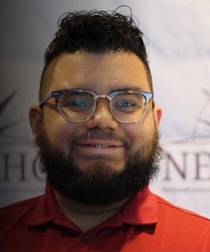 A person with short, curly dark hair, a full beard, glasses, and a red collared shirt smiles in front of a blurred background with partially visible text and shapes.