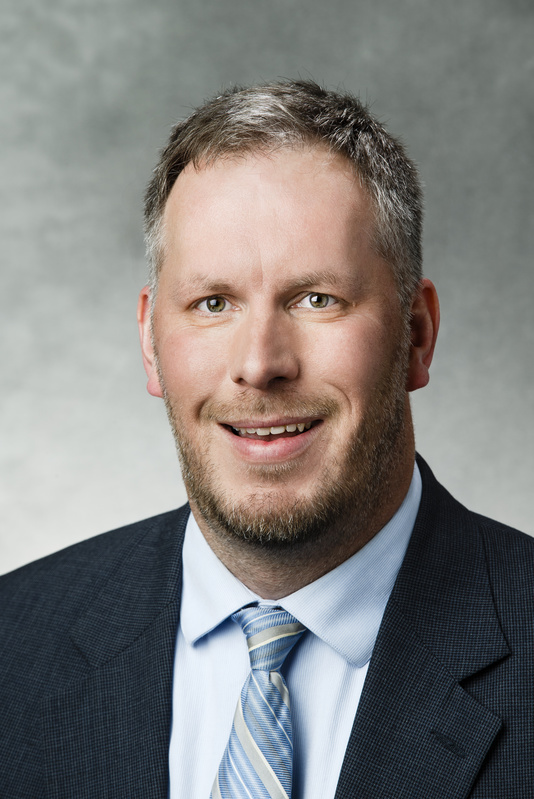 Smiling man with short gray hair and beard, wearing a dark suit, light blue shirt, and striped tie, posing in front of a neutral gray background.