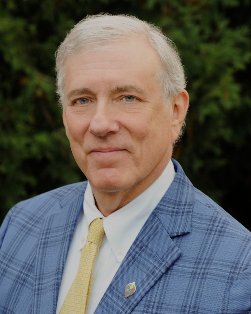 A man with gray hair wearing a blue plaid blazer, white shirt, and yellow tie, standing outdoors in front of green foliage.