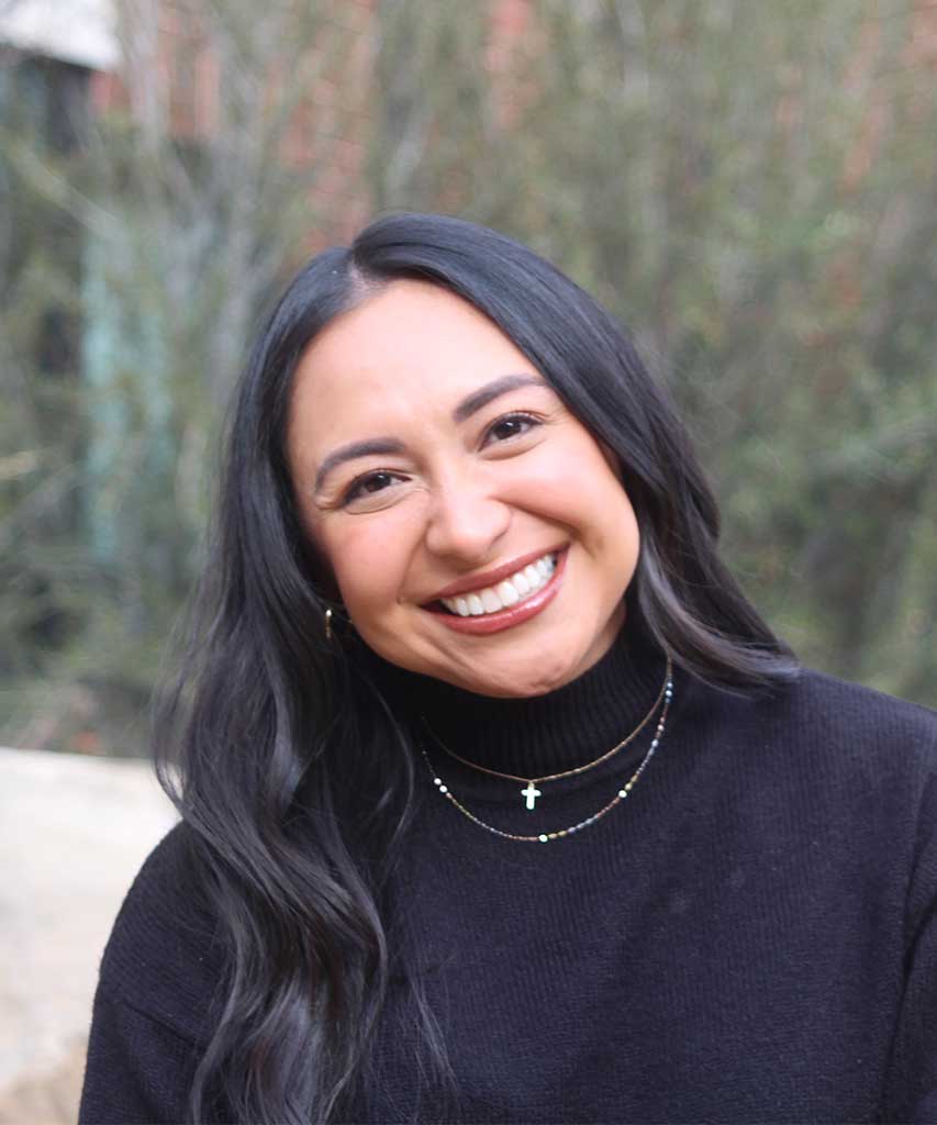 Woman with long dark hair, wearing a black turtleneck and layered necklaces, smiles outdoors with blurred greenery in the background.