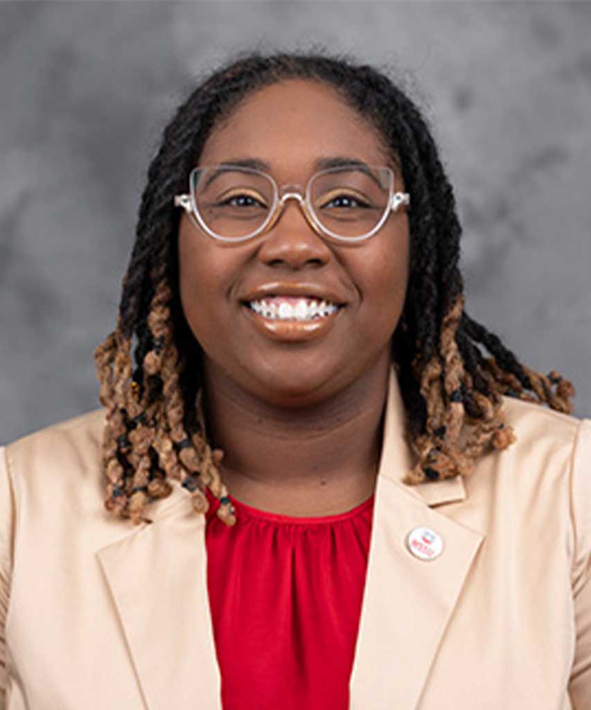 A person with glasses and shoulder-length locs, wearing a beige blazer over a red top, smiling at the camera, with a gray background.