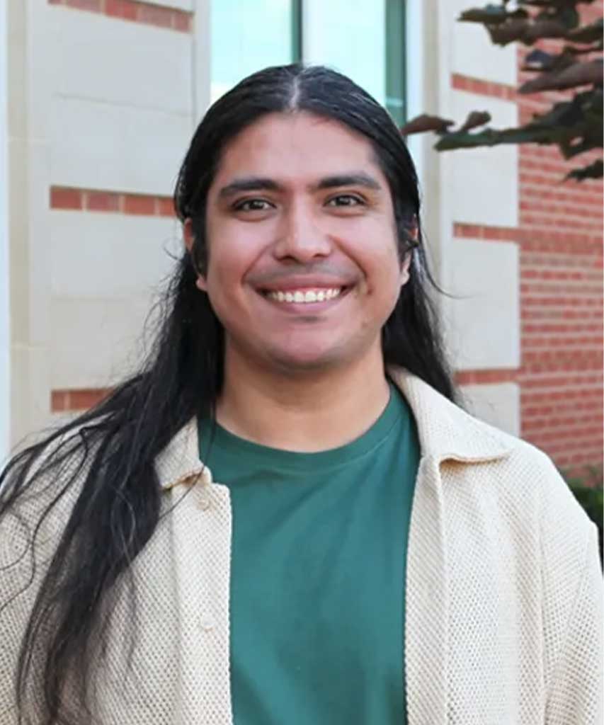 A person with long dark hair smiles outdoors, wearing a green shirt and a light-colored sweater. Brick and stone building walls and greenery are visible in the background.