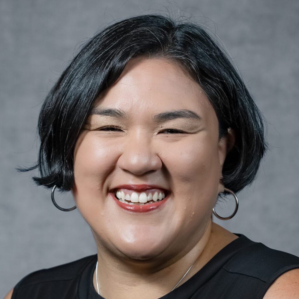 A woman with short black hair and hoop earrings smiles warmly. She is wearing a sleeveless black top and a necklace, with a gray textured background behind her.