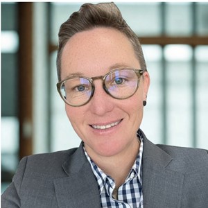 Person with short hair, glasses, and a checked shirt under a gray blazer smiles at the camera in a well-lit, modern office setting with large windows in the background.