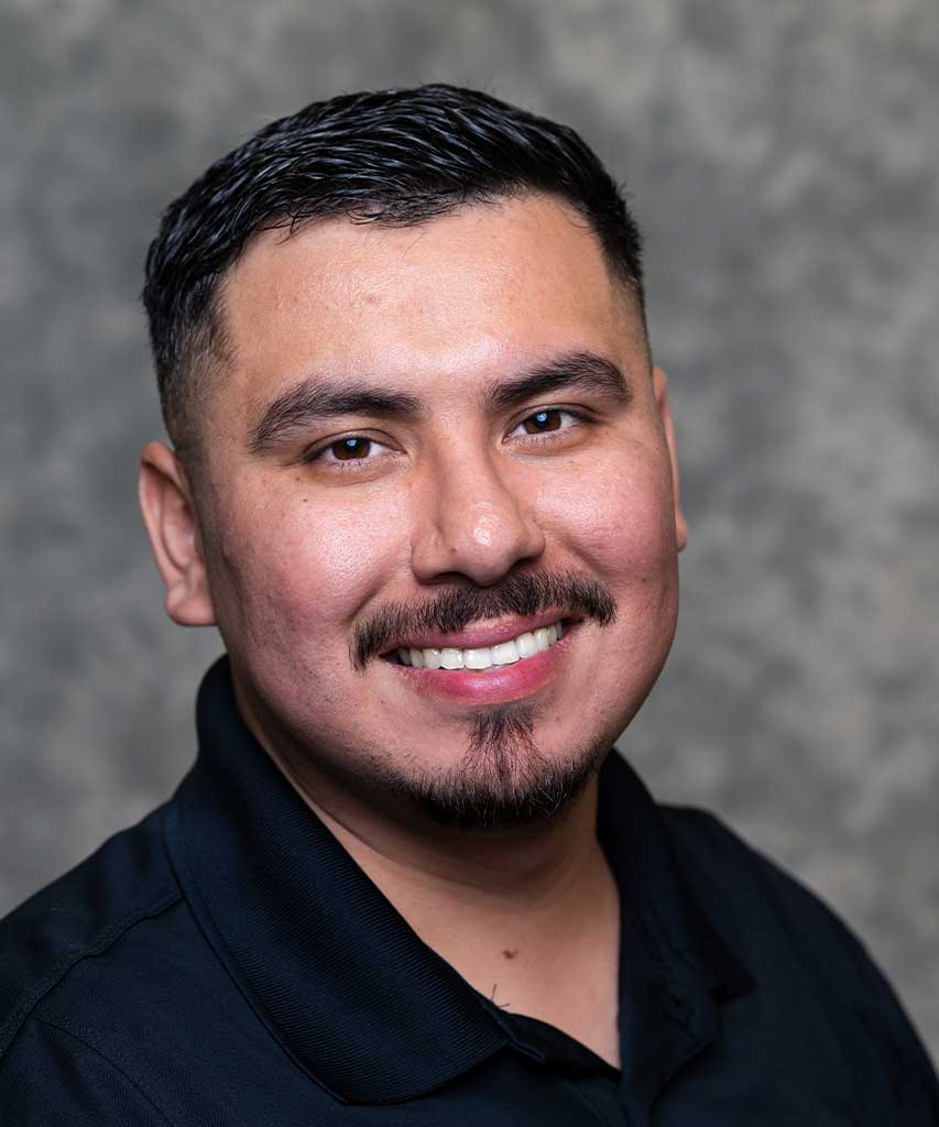 A man with short dark hair and a beard, wearing a black collared shirt, smiles at the camera in front of a gray textured background.