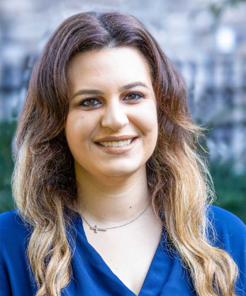 A woman with long wavy brown hair and a blue blouse smiles outdoors. She wears a delicate necklace and stands in front of a blurred, natural background.