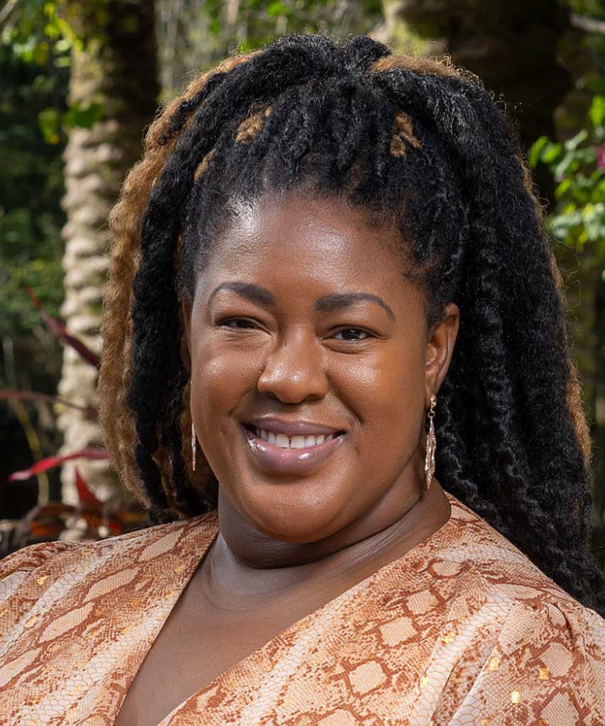 A woman with long, curly black hair smiles outdoors. She wears a light brown patterned top and gold earrings. Trees and greenery are visible in the background.